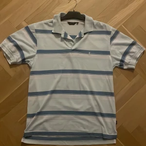 Randig pikétröja från Yves Saint Laurent - Snygg vintage pikétröja från Yves Saint Laurent i ljusblått och vitt med breda blå ränder. Klassisk krage, korta ärmar och broderad. PRIS KAN DISKUTERAS