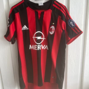 AC Milan Kaka #22 fotbollströja Adidas - Säljer en klassisk AC Milan fotbollströja från Adidas med Kaka och nummer 22 på ryggen. Tröjan är röd och svart med vertikala ränder, klubbmärke och Champions League-märke på ärmen. Tillverkad i lätt och ventilerande material, perfekt för fotbollsälskare.