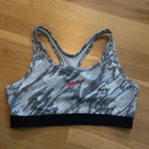 Nike sporttopp gråmönstrad - Sportig grå och svart mönstrad sporttopp från Nike med rosa logga framtill. Tillverkad i Dri-FIT-material som andas och transporterar bort fukt. Brottarrygg och bred resår nedtill för extra stöd. Perfekt för träning och aktiva dagar.