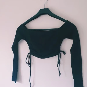 Svart croppad topp från FB Sister - Svart långärmad croppad topp från FB Sister med snörning i sidorna. Toppen har en bred, något fyrkantig halsringning och är tillverkad i ribbad bomull som ger en skön och stretchig känsla. Perfekt för dig som gillar trendiga och stilrena plagg.  Lite ljusare i färgen & små noppror annars fint skick