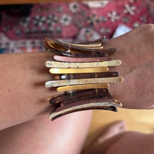 armband i guld och brunt - armband med böjda stavar i guld, beige och olika bruna toner. Armbandet har en cool, modern design med metalliska och marmorerade detaljer som ger en riktigt statement-look. Perfekt för dig som vill sticka ut med accessoarerna.