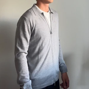 J.Lindeberg Ljusgrå kofta med dragkedja - Snygg ljusgrå J.Lindeberg kofta med hel dragkedja och klassisk krage. Tröjan är stickad i ett mjukt material och har långa ärmar med ribbade muddar. Perfekt för lager-på-lager och passar till många olika stilar.