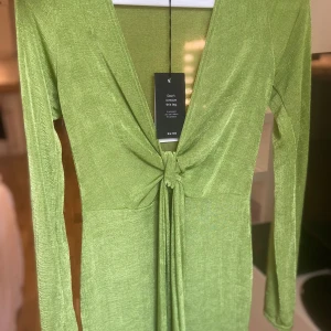 Grön långärmad klänning från NA-KD - Snygg grön klänning från NA-KD med lång ärm och djup v-ringning. Klänningen har en knytning framtill som ger en cool detalj och markerar midjan. Materialet har en lätt glansig finish och känns mjukt mot huden. Perfekt för dig som vill sticka ut med färg.