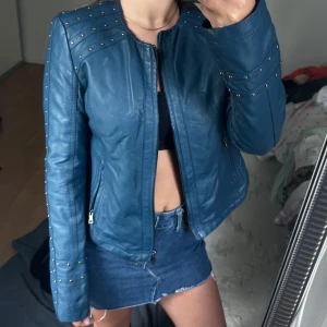 Blå skinnjacka med nitar - Säljer en blå skinnjacka med coola nitar på axlar och ärmar. Jackan har dragkedja framtill och två fickor med dragkedja. Snygg passform och perfekt för att lyfta vilken outfit som helst.