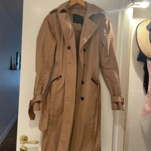 Beige trenchcoat från Zara Basic - Snygg beige trenchcoat från Zara Basic med klassisk krage, knappar framtill och bälte i midjan. Jackan har dragkedjor vid fickorna och detaljer vid ärmsluten. Perfekt för dig som vill ha en stilren och tidlös look. Pytte prickar som inte syns eller stör, går kanske bort i tvätten har ej testat!