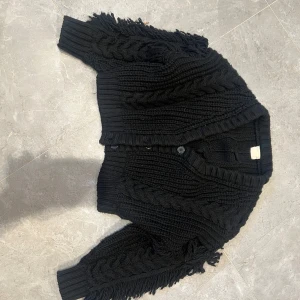 Svart stickad kofta med fransar - Säljer en svart, chunky stickad kofta med grova flätstickade detaljer och fransar längs ärmar och nederkant. Koftan har knappar framtill och en loose, croppad passform som ger en cool och avslappnad vibe. Perfekt att slänga över en t-shirt eller topp.