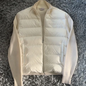 Vit dunväst från Moncler med stickade ärmar - Snygg vit dunväst från Moncler med quiltad framsida och stickade ärmar och rygg. Västen har dragkedja framtill, ribbad krage och diskret Moncler-logga på bröstet. Perfekt för lager-på-lager och ger en clean, exklusiv look.