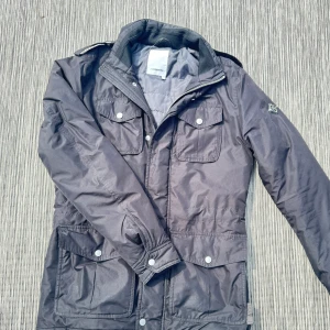 Svart parkajacka från J.Lindeberg - Snygg svart parkajacka från J.Lindeberg med flera praktiska fickor framtill och coola detaljer som axelklaffar och logotyp på ärmen. Jackan har dragkedja och tryckknappar, perfekt för dig som gillar stilren och funktionell design.