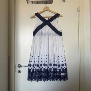 Vit och marinblå sommarklänning med brodyr - Superfin vit kortklänning med marinblå broderade blommor och prickar nertill. Klänningen är ärmlös med breda korslagda band i ryggen och har en markerad midja med marinblå spets. Perfekt för varma dagar och har en lätt, luftig känsla. Från märket Odd Molly i storlek 2 som enligt deras storleksguide motsvarar en storlek M. Fint skick, men tyvärr finns det anmärkning vid armhålan. 