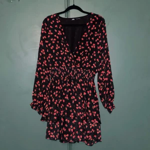 Svart blommig kortklänning från Zara - Supersöt svart kortklänning från Zara med röda blommor och små vita prickar. Klänningen har v-ringning, långa ärmar med resår vid handlederna och markerad midja. Tunt, flowy material som ger en lätt och luftig känsla.
