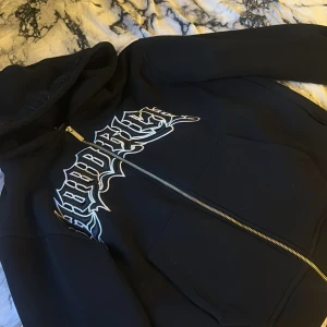Svart hoodie med tryck från Hoodrich - Svart hoodie från Hoodrich i storlek S med stort, vitt och silvrigt tryck på bröstet och broderad text på huvan. Tröjan har dragkedja framtill och två stora fickor. Perfekt för dig som gillar streetwear och vill sticka ut med en fet logga.