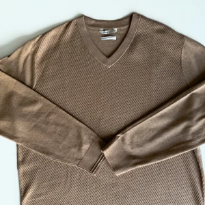 Beige stickad tröja Tom Tailor  - En helt ny stilren, ljusbrun V-ringad tröja med ett subtilt texturerat mönster på framsidan. Tröjan har långa ärmar. 40% Bomull 60% Polyester. Nypris 560 kr.