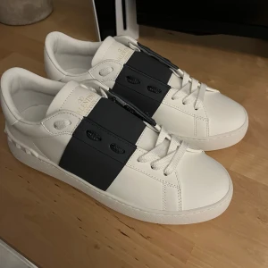 Valentino Open sneakers vit/svart skinn - Snygga Valentino Open sneakers i vitt skinn med bred svart rem över vristen och diskret logga på plösen. Klassisk rund tå, platt sula och coola nitar baktill. Perfekta för dig som gillar stilrena och lyxiga detaljer.