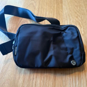Svart axelväska från Lululemon - Snygg svart axelväska från Lululemon med justerbart band och dragkedja. Väskan är tillverkad i nylon med foder av återvunnen polyester och har flera smarta innerfack. Perfekt storlek för att bära med sig det viktigaste och passar till många olika outfits. Helt ny