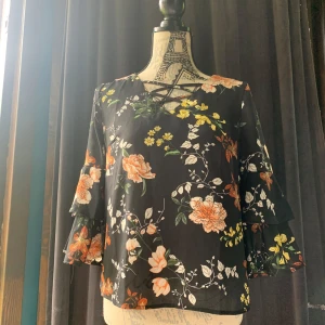 Blommig blus med volangärm  - Supersöt svart blus från Amisu med färgglada blommor i gult, orange och vitt. Blusen har v-ringning med snörning och volangärmar i trekvartsmodell. Luftigt material som känns lätt och flowy, perfekt för dig som gillar romantiska vibes.