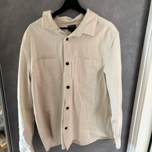 Beige overshirt i manchester - Stilren beige overshirt från hm. Skjortan har två stora bröstfickor, svarta knappar och klassisk krage. Perfekt att bära öppen eller stängd för en avslappnad look. Långärmad och med en lite loose passform.