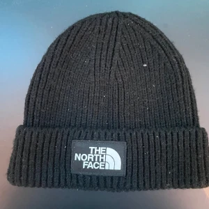 Svart ribbstickad mössa The North Face - Svart ribbstickad mössa från The North Face med klassisk uppvikt kant och tydlig logotyp framtill. Tillverkad i mjuk akrylblandning, perfekt för kalla dagar. Unisexmodell som passar de flesta huvudstorlekar.