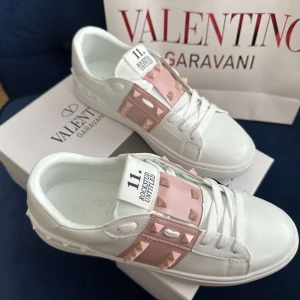 Valentino Rockstud sneakers vit/rosa - Snygga Valentino Rockstud sneakers i vitt skinn med ljusrosa band och ikoniska nitar. Klassisk rund tå, platt sula och coola detaljer med nitar runt hälen och på sidan. Perfekta för dig som vill sticka ut med exklusiv streetstyle.