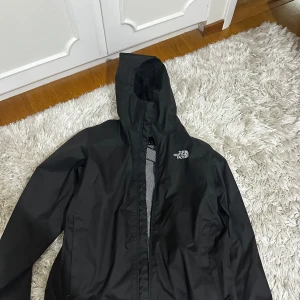 Svart vindjacka från The North Face - Svart vindjacka från The North Face med huva och dragkedja framtill. Jackan har meshfoder på insidan för extra ventilation och en stilren look med logga på bröstet. Perfekt för blåsiga dagar och enkel att matcha med allt.