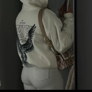 Vit hoodie med vingar och tryck - Säljer en vit hoodie med stor svart vinggrafik och text på ryggen. Tröjan har huva och en avslappnad passform, perfekt för en cool och avslappnad stil. Materialet känns mjukt och skönt mot huden.