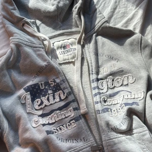 Grå hoodie från Lexington  - Snygg grå hoodie från Lexington med broderad logga och tryck framtill. Tröjan har huva med snörning, dragkedja och kängurufickor. Mjuk bomullskänsla och långärmad modell.