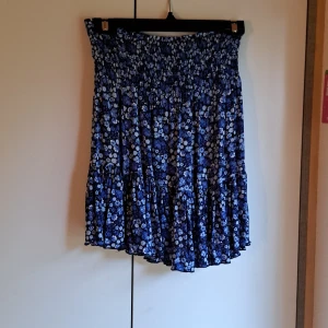 Blå blommig volangkjol - Supersöt blå kortkjol med vita blommor och volangkant. Kjolen har smock i midjan för en bekväm passform och är perfekt för varma dagar. Lätt och luftig känsla, passar perfekt till sneakers eller sandaler.