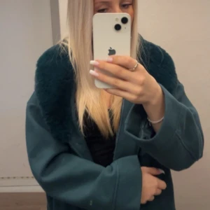 Mörkgrön jacka med fuskpäls-krage - Snygg mörkgrön jacka med stor, mjuk fuskpäls-krage. Jackan har två stora fickor framtill med lock och knappar. Modellen är boxig och lite oversized, perfekt för dig som vill sticka ut med en cool look. Materialet känns tjockt och värmande.