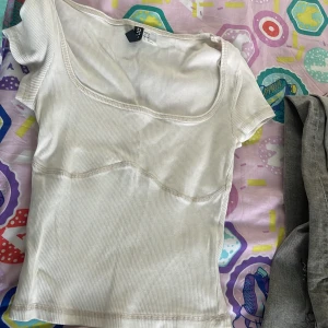 Ribbad vit topp från Divided XS - Snygg ribbad topp från Divided i vit färg med korta ärmar och markerad söm under bysten. Toppen har en bred, rundad urringning och är i stretchigt material som sitter tajt. Perfekt att styla med jeans eller kjol för en clean look.