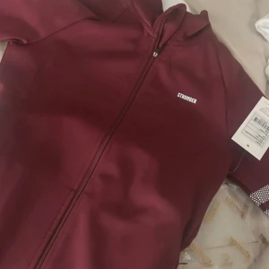 Vinröd träningsjacka från Stronger S - Vinröd träningsjacka från Stronger med dragkedja framtill och vita detaljer på ena ärmen. Jackan är figurnära, har lång ärm och är tillverkad i stretchigt material (92% polyester, 8% elastan).helt ny och aldrig andvänd! Perfekt för dig som vill ha en snygg och sportig look.