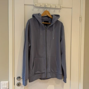 Blå hoodie med dragkedja från 157 - Mysig blå hoodie från 157 i storlek L. Tröjan har huva med snörning, hel dragkedja och två stora fickor framtill. Tillverkad i mjuk bomullsmix, perfekt för chill dagar. Enkel och stilren design som funkar året runt.