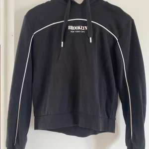 Svart hoodie från H&M - Svart hoodie från Divided i storlek XS med vit text 'BROOKLYN NEW YORK CITY' på bröstet och vita detaljer längs ärmar och bröst. Klassisk huva med snörning och långa ärmar. Perfekt för en chill och urban look.