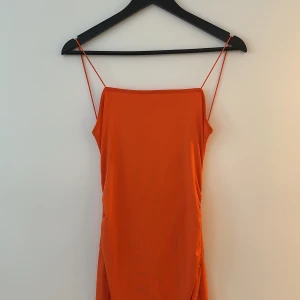 Orange långklänning med smala band - Snygg orange långklänning med smala axelband och rak urringning. Klänningen har en slits på sidan och är tillverkad i ett mjukt, lätt material som faller fint. Perfekt för dig som vill sticka ut med en färgstark look.