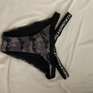 Camouflage bikiniunderdel med textband - Snygg bikiniunderdel med grått och svart camouflage-mönster. Breda svarta resårband med vit text där det står #LONDON och #TOKYO ger en cool streetstyle-vibe. Perfekt för stranden eller poolen om du vill sticka ut.