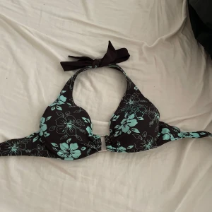 Blommig bikiniöverdel med turkosa detaljer - Snygg bikiniöverdel med svart botten och stora turkosa blommor. Dekorerad med små turkosa stenar på ena kupan för extra bling. Halterneck-modell med knyt i nacken och vadderade kupor. Perfekt för stranden eller poolen!