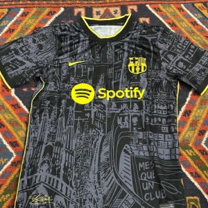 Svart och gul FC Barcelona fotbollströja - Snygg FC Barcelona tröja i svart med detaljer i gult. Tröjan har ett unikt mönster med stadsmotiv och klubbens emblem på bröstet. Tillverkad i Dri-FIT material för optimal komfort. Perfekt för fans som vill sticka ut på läktaren!