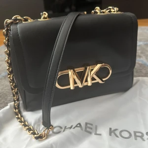 Svart axelväska från Michael Kors - Snygg svart axelväska från Michael Kors med guldfärgade detaljer och kedjerem. Väskan har ett stort MK-spänne framtill och innerfack med dragkedja. Tillverkad i slätt skinn och perfekt för dig som gillar stilrena accessoarer.