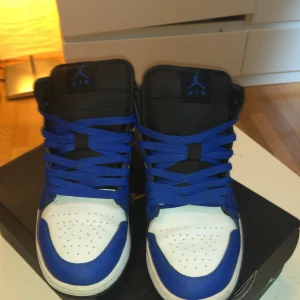Nike Air Jordan 1 blå och vita sneakers - Säljer ett par Nike Air Jordan 1 sneakers i blått, vitt och svart. Skorna har klassisk Jordan-logga på sidan, svarta swoosh och blå snörning. Ovandelen är i skinn med perforerad tåbox och svart plös. Perfekta för dig som gillar streetwear och vill sticka ut.