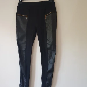 Svarta leggings med skinnpaneler Favolook - Svarta leggings från Favolook med snygga skinnimitation-paneler framtill och guldfärgade dragkedjor vid fickorna. Tight passform och coola detaljer som ger en edgy vibe. Perfekta för dig som gillar streetstyle och vill sticka ut.  Detalj fickor bak & fram Trådsläpp (se extra bild) & noppror annars fint skick