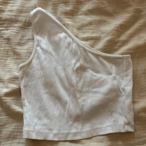 Vit oneshoulder topp från Even&Odd - Säljer en vit ribbad oneshoulder topp från Even&Odd. Toppen är ärmlös med asymmetrisk skärning och sitter tight. Perfekt för dig som gillar minimalistisk stil och vill ha något som sticker ut lite extra.