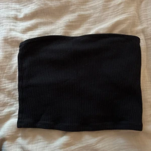 Svart ribbad bandeau tubtopp - En svart bandeau tubtopp med ribbad struktur. Toppen är axelbandslös och har en enkel, stilren design som passar perfekt till jeans eller kjol. Mjuk och stretchig för en bekväm passform.