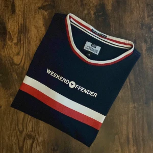 Weekend Offender T-Shirt - Weekend Offender t-shirt i navy med röd och vit rand samt tryck. Storlek S. Klassisk brittisk casual-stil med “Too Much Too Young” etikett i kragen. Perfekt till jeans eller shorts – en ikonisk streetwear-look. I mycket bra skick, inga fläckar eller hål.