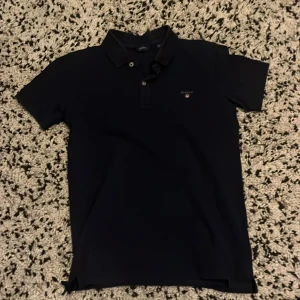 Svart pikétröja från GANT - Klassisk svart pikétröja från GANT med broderad logga på bröstet. Tröjan har krage, korta ärmar och knäppning med två knappar framtill. Perfekt för en clean och stilren look. Den är lite stor i storlek så skulle säga att den skulle passa de som är 155-162