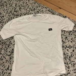 Vit t-shirt från C.P. Company - Vit t-shirt från C.P. Company med klassisk passform och korta ärmar. T-shirten har en liten svart patch med logga på bröstet och är tillverkad i mjukt material som känns skönt mot huden. Perfekt för en clean och stilren look. Har bara använt den 2 gånger.