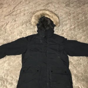 Svart dunjacka med fuskpäls från Everest - Svart dunjacka från Everest med huva och fuskpäls. Jackan har långa ärmar, dragkedja och knappar framtill samt stora fickor. Insidan är quiltad för extra värme och det finns reflexdetaljer på ärm och rygg. Perfekt för kalla vinterdagar. Jag köpte den jackan ett tag sen men har inte använt den så mycket.Jackan är i bra skikt.storleken är i 158 men om du har storleken M så kommer den 100% funka på dig