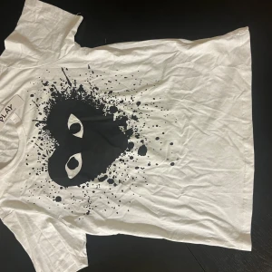 Vit t-shirt  - Vit t-shirt från med ett stort svart tryck framtill som föreställer ett stänkmönster med ögon. Klassisk passform och rund hals. Tillverkad i mjuk bomull, perfekt för en clean och cool streetstyle. OBS!!! INTE ÄKTA