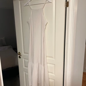 Ljus långklänning från 157 - Säljer en ljus, ärmlös långklänning från 157 med tunna axelband och volang nedtill. Klänningen har en enkel, flowy passform och är tillverkad i ett strukturerat tyg som känns luftigt och bekvämt. Perfekt för varma dagar och somriga vibes.