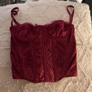 Vinröd spetskorsett  - Supersnygg vinröd korsett med spets och genomskinliga meshpaneler. Justerbara axelband, dekorativa sömmar och hakknäppning bak. förliten för mig så har inga bilder med den på tyvärr.