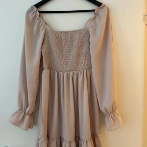 Beige långärmad klänning med volang - Supersöt beige klänning med långa puffärmar och smockad överdel. Klänningen har volangdetaljer nertill och en luftig, flowy passform. Perfekt för dig som gillar romantisk och trendig stil.
