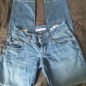 Blå straight jeans från Pepe Jeans - Säljer ett par klassiska blå jeans från Pepe Jeans i modellen Venus. De har låg midja, raka ben och snygga detaljer med kontrastsömmar. Jeansen är tillverkade i bomull och har en stilren look med fem fickor och läderpatch bak.