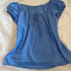 Blå blus med knyt och knappar - Supersöt blå blus med kort puffärm, rundad halsringning och knytband framtill. Dekorativa knappar och veckade detaljer vid bysten ger en romantisk vibe. 🫶🏻🫶🏻